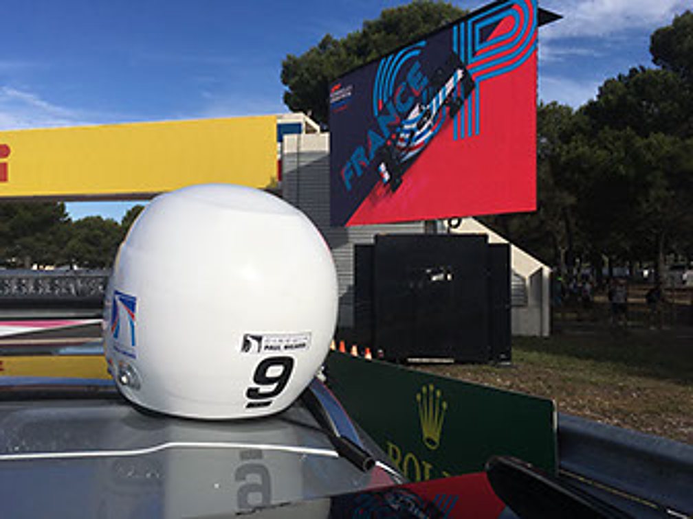 Un casque blanc portant le numéro 9 est posé sur une voiture de course, avec un grand écran rouge et des arbres en arrière-plan.