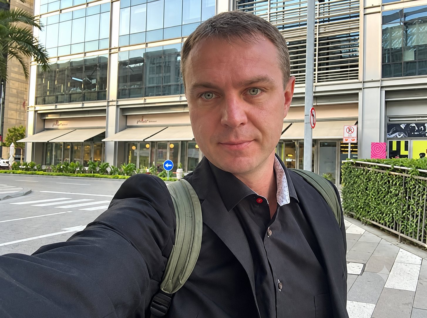 Un homme aux cheveux courts portant une chemise sombre et un sac à dos prend un selfie dans la rue devant un immeuble moderne à façade vitrée.