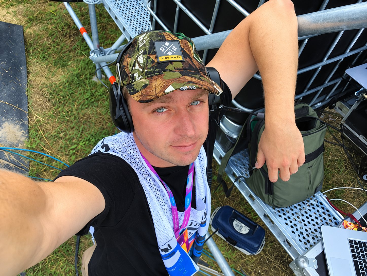 Un homme portant une casquette camouflage et un casque audio se prend en photo en hauteur sur une structure métallique, sur un site en plein air.