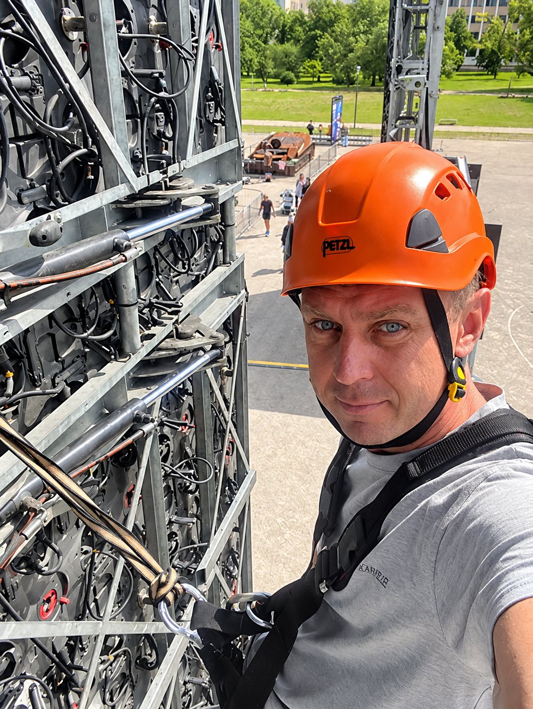 Un technicien portant un casque orange et un harnais prend un selfie devant une structure métallique couverte de câbles, avec un parc en arrière-plan.
